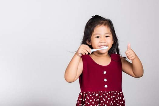 Little cute kid girl 3-4 years old smile brushing teeth and show thumb up finger for good sign in studio shot isolated on white background, happy Asian children, Dental hygiene healthy concept