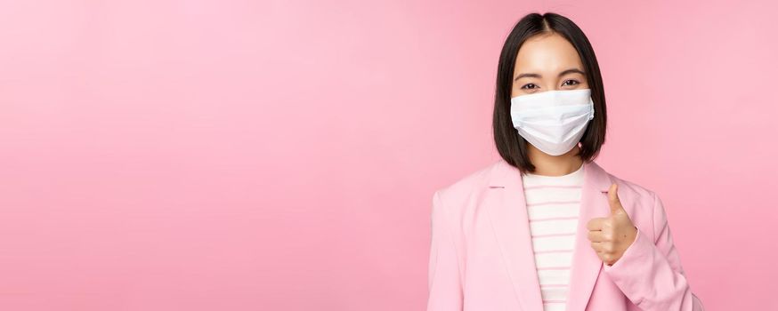 Asian businesswoman in suit and medical face mask, showing thumbs up, recommending wearing personal protective equipment in office during covid-19 pandemic, pink background.