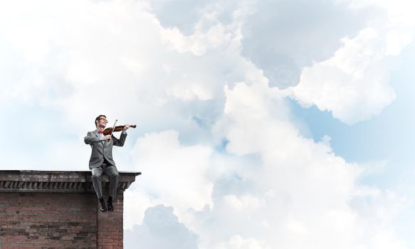 Young man wearing suit and glasses sitting on roof and playing violin