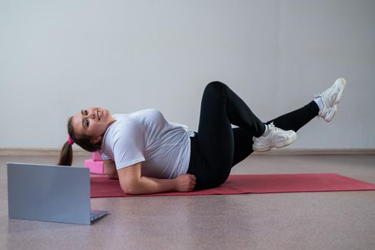 A chubby young woman is watching an online fitness lesson on a laptop. Distance sports training