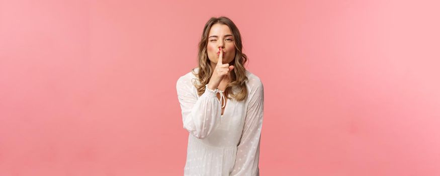 Beauty, fashion and women concept. Portrait of feminine and flirty caucasian woman hiding secret, asking keep silent, shushing with sensual coquettish smile, wear white dress, pink background.