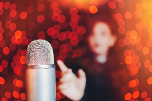 Woman pulls hand to disco mic for sing at concert. Close-up of retro microphone and silhouette of female singer on bokeh blur background.