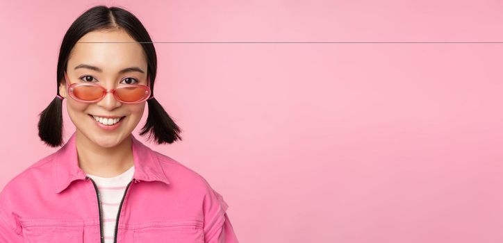 Close up of stylish korean girl in sunglasses, smiling happy, posing against pink background. People face concept.