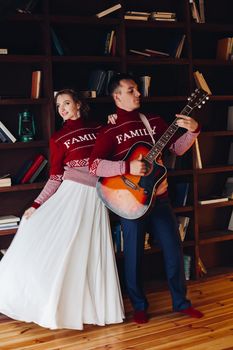 Beautiful smiling couple in red sweaters listening music, girlfriend leaning on handsome boyfriend, when he playing at gitar. Celebrating new year and xmas. Romantic winter holidays.