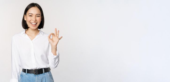 Very good. Smiling asian woman showing okay with satisfied face expression, praise and compliment great job, pleased by smth, standing over white background.