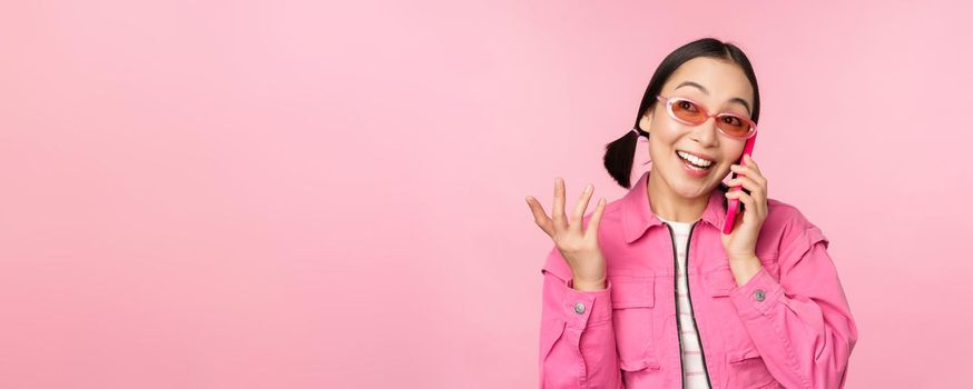 Beautiful korean female model in sunglasses, talking on mobile phone with happy face, using cellular service to call friend on smartphone, standing over pink background.