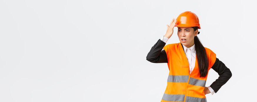 Annoyed and disappointed female asian chief engineer scolding construction workers making stupid mistake, tap helmet and looking bothered, mad at architect, standing white background.
