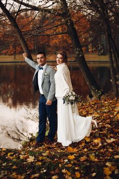 Back view of beautiful young bride in sweater and dress and groom in black coat and suite standing at river or lake pear, embracing and kissing. Beloved couple walking, celebrating wedding at autumn.