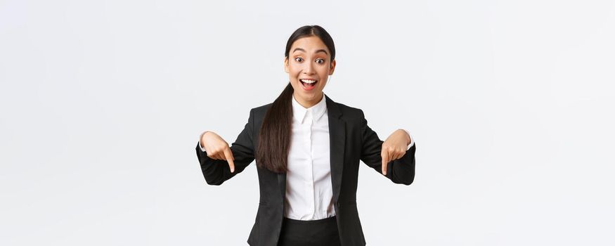 Happy excited asian female entrepreneur in suit pointing fingers down to show best deal, price offer. Industrial woman formal clothes showing bottom advertisement with amused smile, white background.