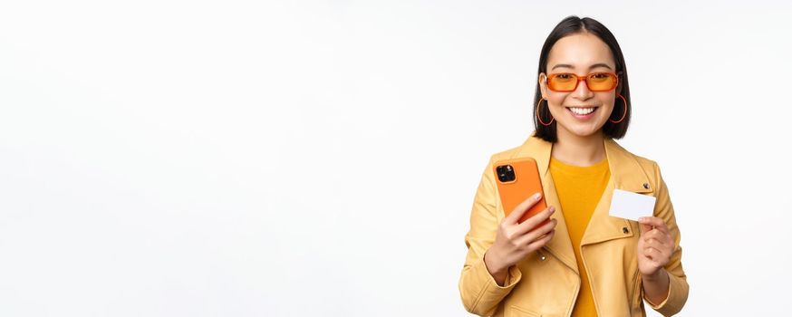 Online shopping. Stylish asian female model in sunglasses, holding credit card and mobile phone, smiling happy, standing over white background. Copy space