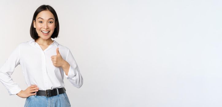 Very good. Smiling enthusiastic asian female entrepreneur, showing thumb up in approval, praise excellent job, satisfied with results, standing over white background.