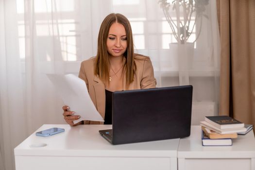 European professional woman sitting with laptop at home office desk, positive woman studying while working on PC. She is wearing a beige jacket and jeans