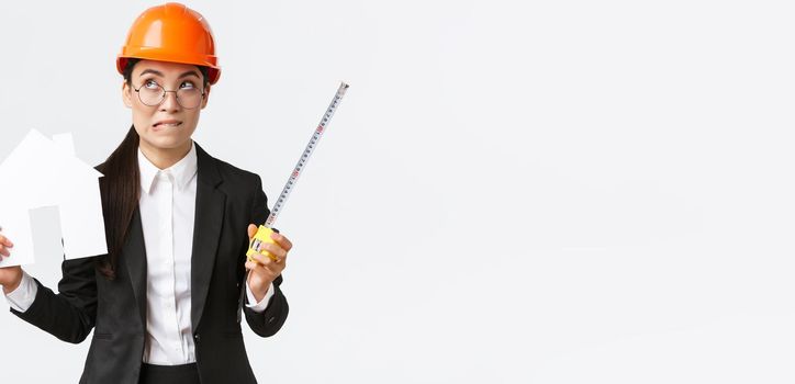Indecisive asian female architect, construction engineer in helmet and business suit holding house maket and tape measure, biting lip unsure, looking away thoughtful, searching solution.