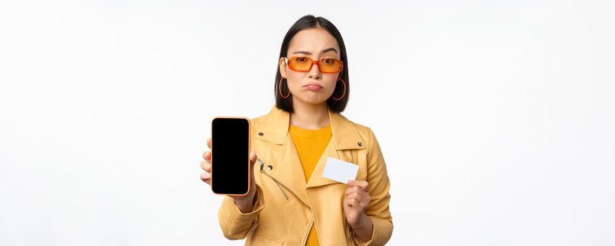 Sad asian girl in sunglasses, showing smartphone app interface, credit card, looking disappointed, standing over white background. Copy space
