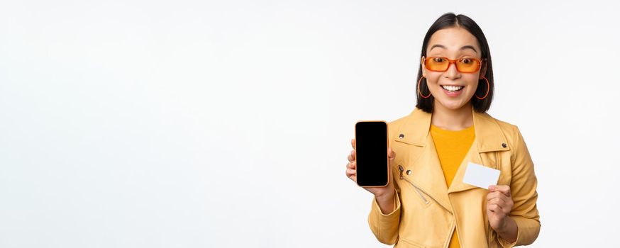 Online shopping and people concept. Stylish asian woman showing mobile phone screen and credit card, smartphone application, standing over white background. Copy space