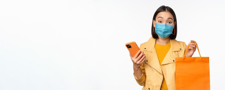 Image of stylish asian girl shopper, holding smartphone and shopping bag without store logo, wearing medical face mask from covid-19, white background.