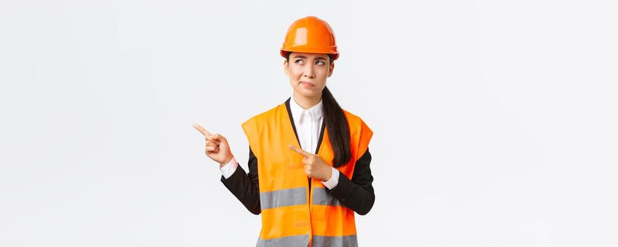 Building, construction and industrial concept. Indecisive asian female architect being unsure, wearing reflective clothing and helmer, smirk as pointing and looking upper left corner puzzled.