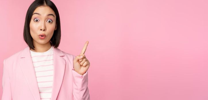 Got an idea. Young asian saleswoman, office manager raising finger, suggesting, wearing suit, posing against pink studio background.