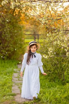 A pretty girl in a white dress and a hat walks under flowering trees in the springs garden