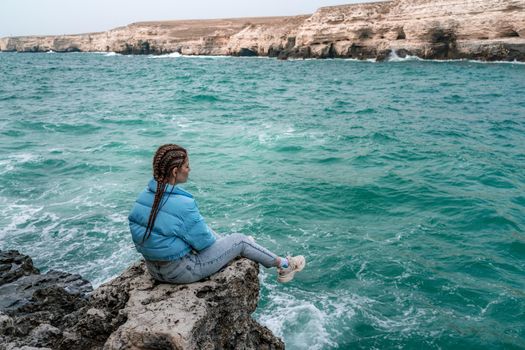 A woman in a blue jacket sits on a rock above a cliff above the sea, looking at the stormy ocean. Girl traveler rests, thinks, dreams, enjoys nature. Peace and calm landscape, windy weather