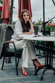 Smiling young dark-haired businesswoman makes notes in a notebook in a cafe on a summer day. Business, e-learning, freelance concept. laptop on a table.