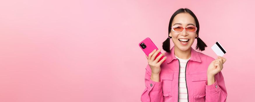 Modern beautiful asian girl, laughing and smiling with mobile phone, credit card, shopping online, paying with smartphone, standing over pink background.
