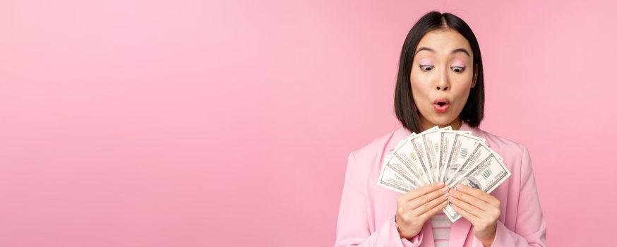 Finance, microcredit and people concept. Happy smiling asian businesswoman showing dollars money, standing in suit against pink background.