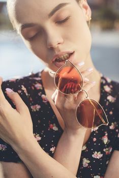 Portrait of a stylish young woman. Brunette with ballerina hairstyle in sundress posing on street, fountain on a background. Sunglasses near lips