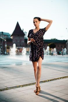 Portrait of a stylish young woman. Brunette with ballerina hairstyle in sundress posing on street, fountain on a background