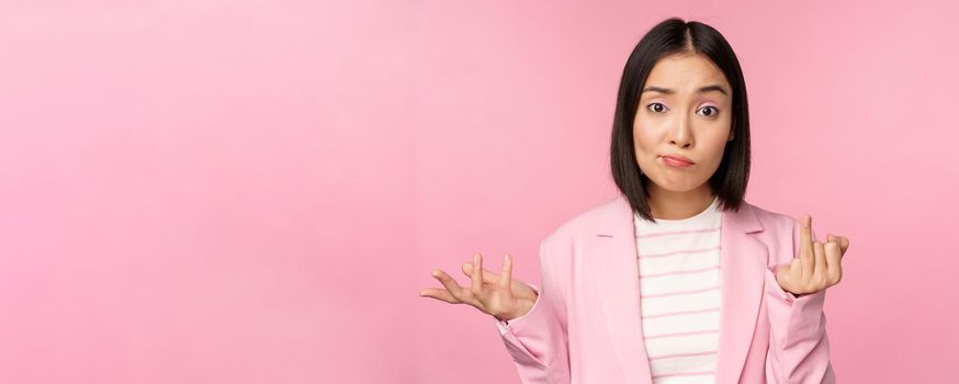 Portrait of young asian business woman, saleswoman shrugging shoulders and looking confused, clueless of smth, standing over pink background.