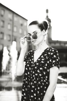 Portrait of a stylish young woman. Brunette with ballerina hairstyle in sundress posing on street, fountain on a background