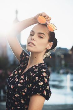 Portrait of a stylish young woman. Brunette with ballerina hairstyle in sundress posing on street, fountain on a background. Sunglasses near lips