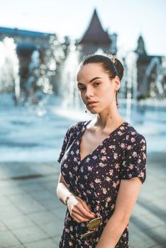 Portrait of a stylish young woman. Brunette with ballerina hairstyle in sundress posing on street, fountain on a background