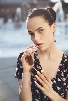 Portrait of a stylish young woman. Brunette with ballerina hairstyle in sundress posing on street, fountain on a background. Sunglasses near lips