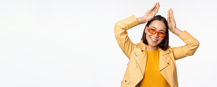 Portrait of beautiful asian girl in sunglasses, showing bunny ears hands gesture and smiling, standing happy over white background.