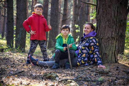 Family with mother and two children boy sibling brothers sits in the forest on felled logs or grass in summer. Happy family in colorful clothes have a hike in autumn forest.