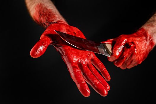 A man with blood-stained hands cuts his palm with a knife on a black background