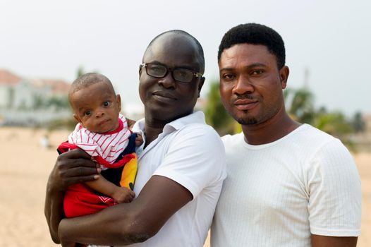 a young man in glasses and dressed in white shirt standing with his friend at the beach holding his baby sticking to his chest and look at the camera.