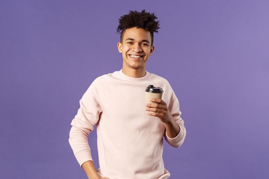 Lifestyle, cafe, eating-out concept. Portrait of cheerful young hispanic guy holding take-away cup of coffee, drinking and smiling camera, waiting for someone, thanking coworker for bringing cappucino.