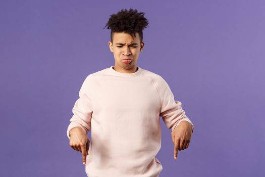 Portrait of gloomy and silly disappointed whining guy, sobbing and crying from envy or jealousy, regret missing chance, looking and pointing down with upset unhappy expression, purple background.