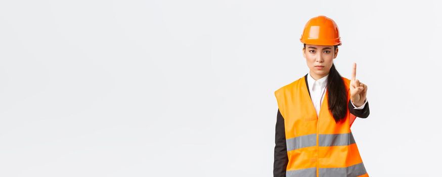 Serious-looking confident and displeased asian female engineer shaking finger, scolding construction worker for acting dangerous, prohibit and forbid action at building zone, white background.