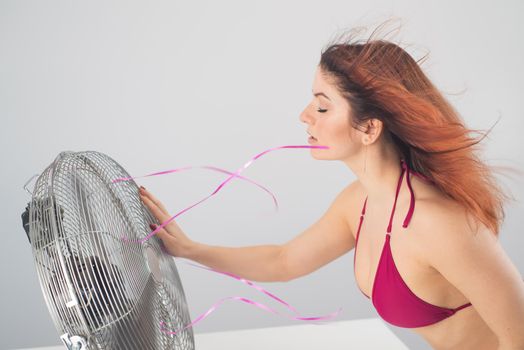 Red-haired smiling woman in a bikini drinks a cold drink and enjoys the blowing wind from an electric fan on a white background. Climate control on a hot summer day.