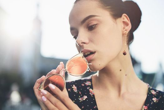 Portrait of a stylish young woman. Brunette with ballerina hairstyle in sundress posing on street, fountain on a background. Sunglasses near lips