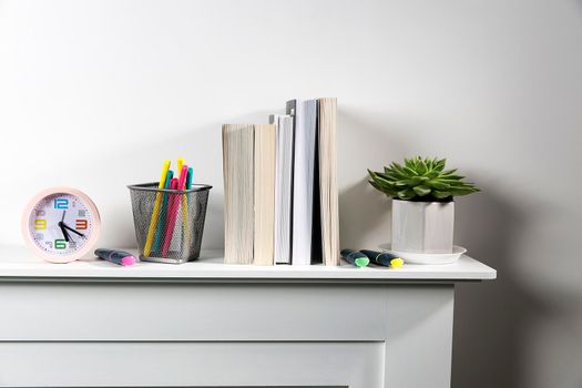 Echeveria in geometric pot, stack of books, pens in a pencil holder, felt-tip pens, are on shelf. Place for text