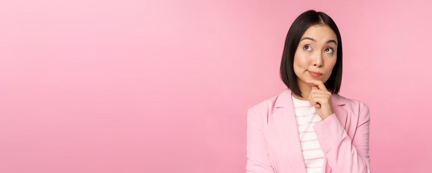 Image of asian businesswoman standing in thinking pose, brainstoming, wearing suit. Korean saleswoman, entrepreneur posing against pink background.