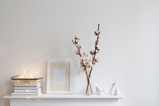 Scandinavian style room interior in white tones. A vase with cotton flowers, a stack of books, a photo frame, a bird figurine on a wooden surface of a shelf. Copy space.