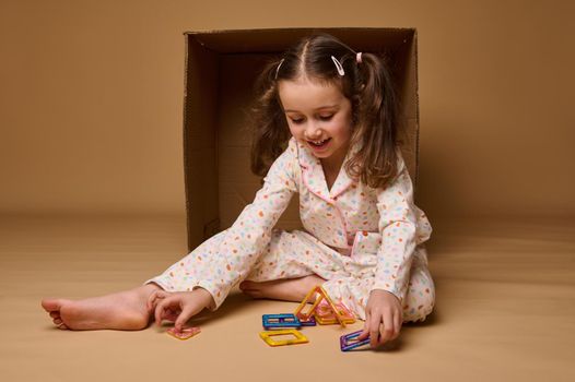 Cute little girl with two ponytails, wearing a colorful pajamas sitting ahead a cardboard box and building a house using square and triangle magnetic construction blocks. Fine motor skills development