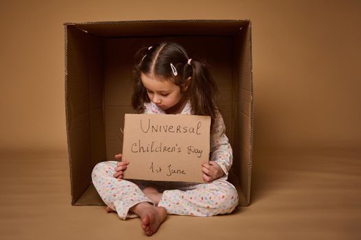 Cute girl with two ponytails holding a cardboard poster sitting inside a cardboard box. International Children's Day. Social advertising, information campaign for the fight for children's rights