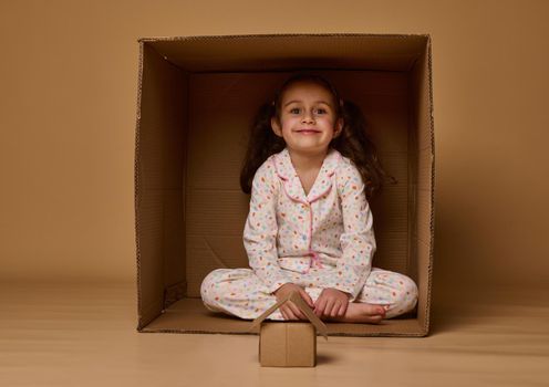 Adorable little European girl sitting inside a cardboard box with a craft house model, smiles looking at camera, posing over beige background, copy ad space. Concept of housing, insurance, real estate