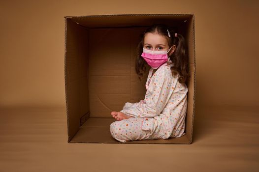 Adorable little European girl with two ponytails in pajamas wearing a pink medical protective mask sits inside a cardboard box, looks at camera, isolated over beige background with copy ad space